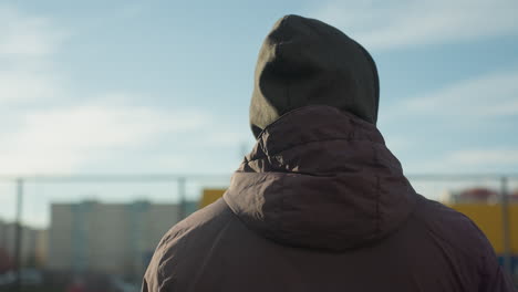 vista trasera de un hombre con capucha y chaqueta mirando a su alrededor en un escenario deportivo urbano con un fondo borroso de edificios residenciales, paredes amarillas vibrantes, iluminadas por la brillante luz del sol