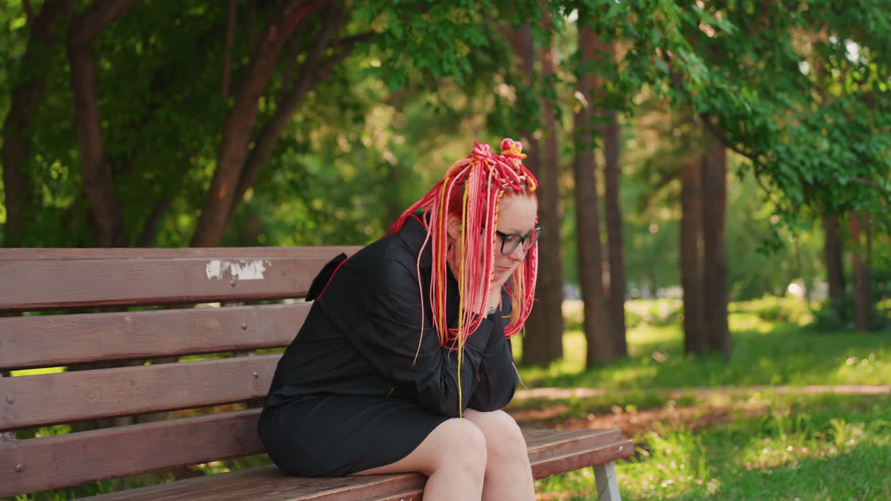 Mujer expresa preocupación, mujer sentada en un parque a la sombra, mujer con cabello brillante y gafas muestra emoción de ansiedad, primer plano de mujer preocupada con rastas vibrantes y gafas en un parque