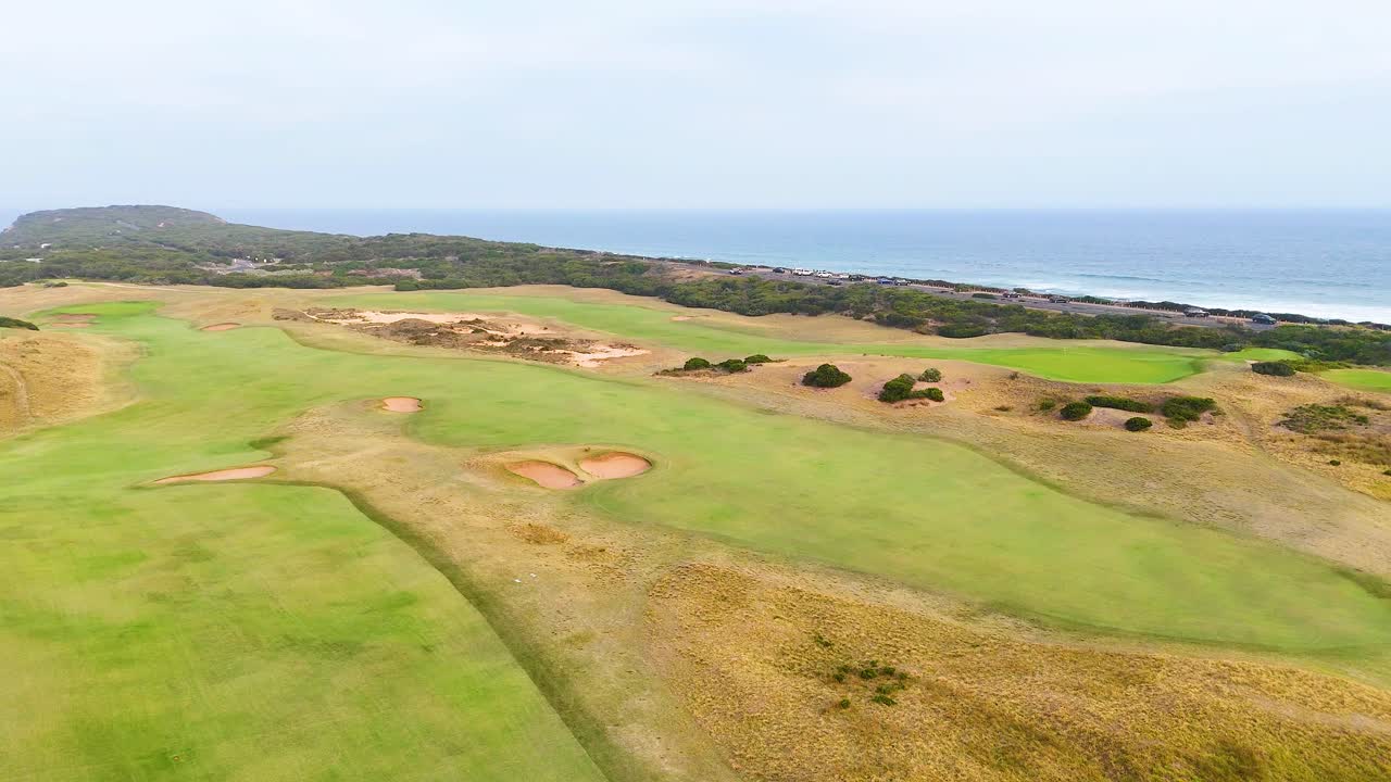 Drone footage captures expansive golf course with ocean backdrop, showcasing lush greens and sandy bunkers under soft daylight
