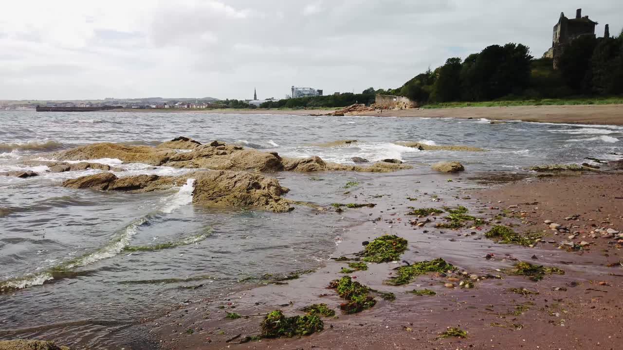 playa dysart en fife escocia con un castillo en el fondo, rompiendo olas, arena y rocas en primer plano