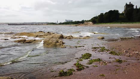 Dysart-beach-in-Fife-Scotland-with-a-castle-in-the-background,-breaking-waves,-sand,-and-rocks-in-the-foreground