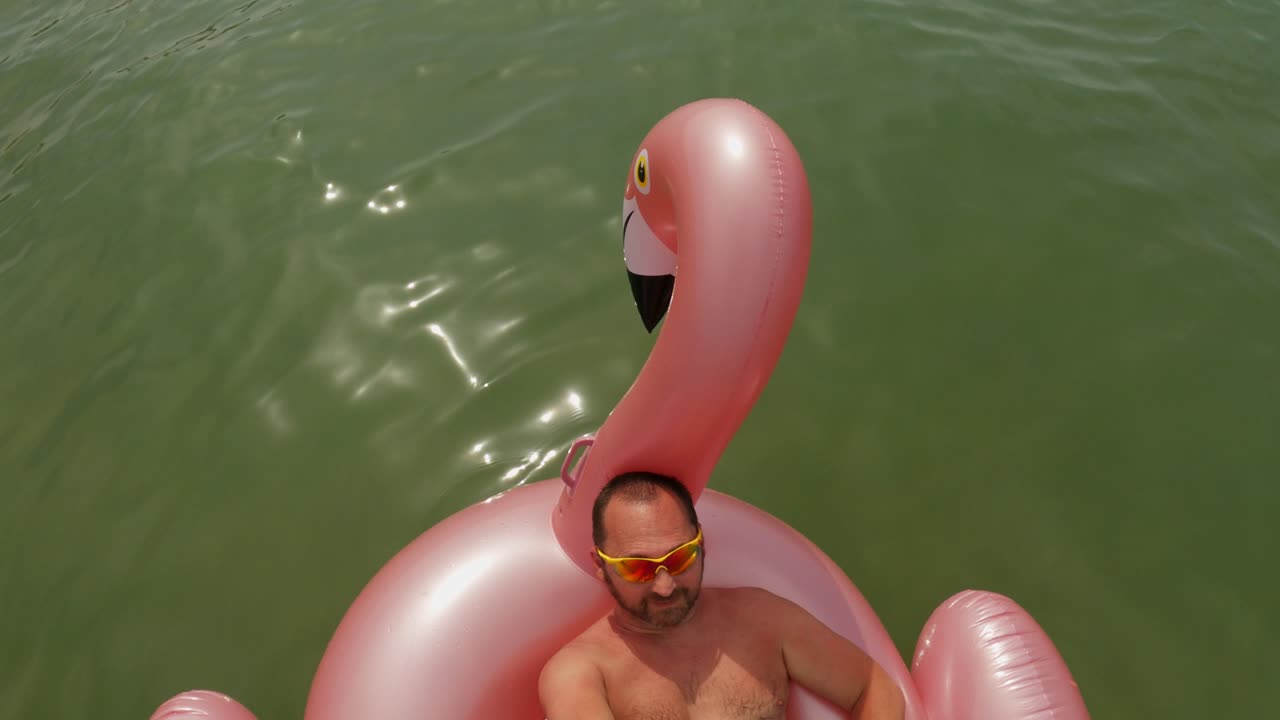 Man with yellow sunglasses taking selfie holding long stick and camera while relaxing on floating inflatable pink flamingo
