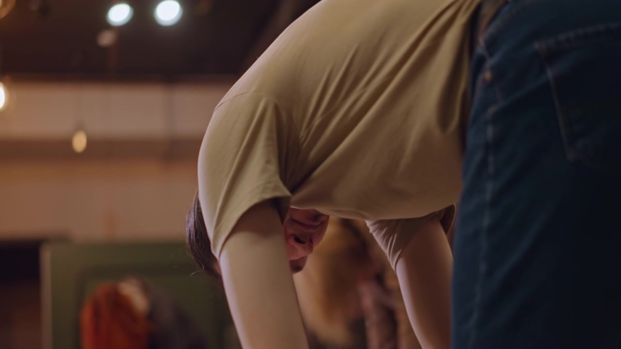 Solo dancer stretching in music studio, intense focus on arm and torso extensions, ceiling lights and mirror reflections, instructor observing posture, slow controlled reach and bend, repetitive