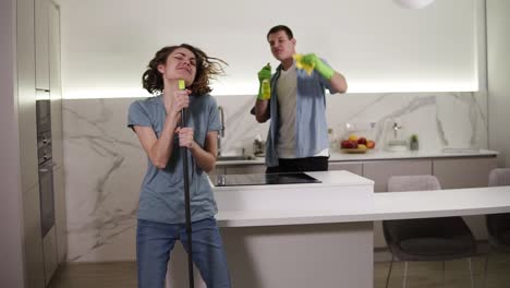 dos jóvenes bailando juntos y cantando emocionalmente usando escoba y herramientas de limpieza como micrófonos mientras limpian la cocina. pareja feliz disfrutando del tiempo. cámara lenta