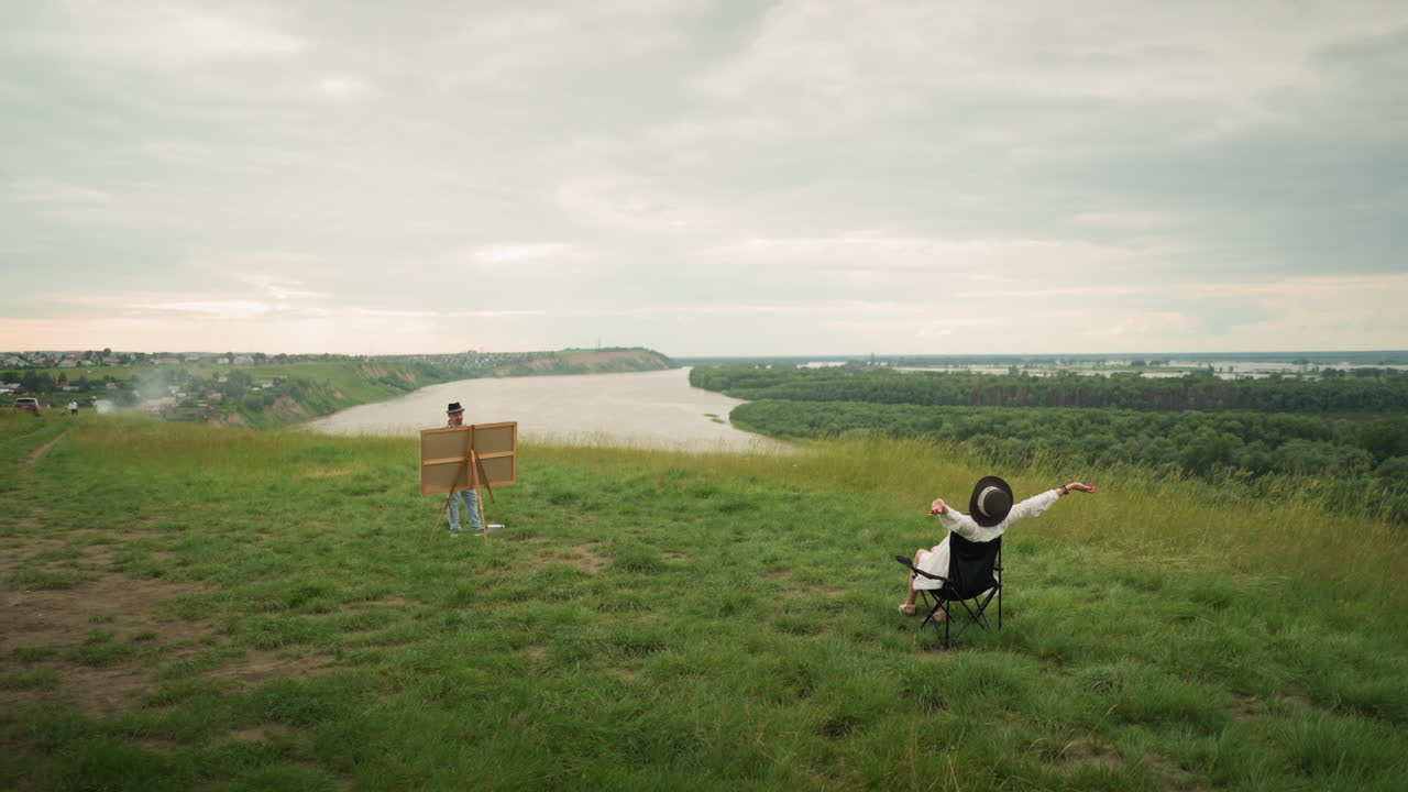 A woman in a black hat sitting comfortably on a chair, stretching her arms while an artist paints her in a lush grass field beside a tranquil lake
