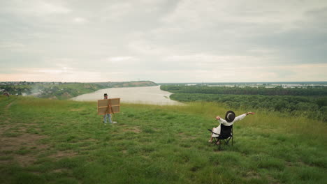 a woman in a black hat sitting comfortably on a chair, stretching her arms while an artist paints her in a lush grass field beside a tranquil lake
