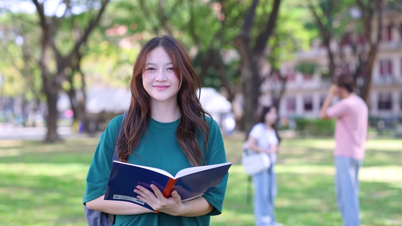Uno studente universitario thailandese legge un libro all'aperto nel campus prima di sorridere alla camera.