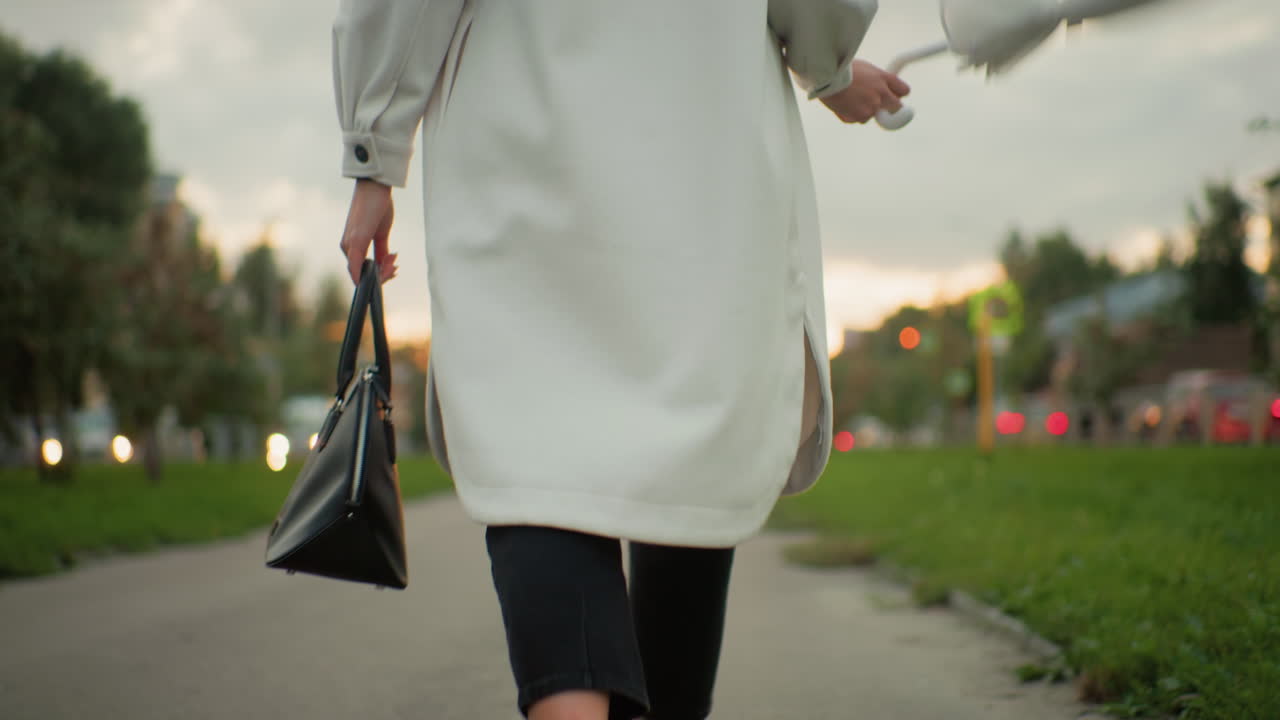 Back view of business woman wearing light coat playfully twirling closed umbrella while walking along paved path bordered by green grass with blurred city lights and moving cars in soft background