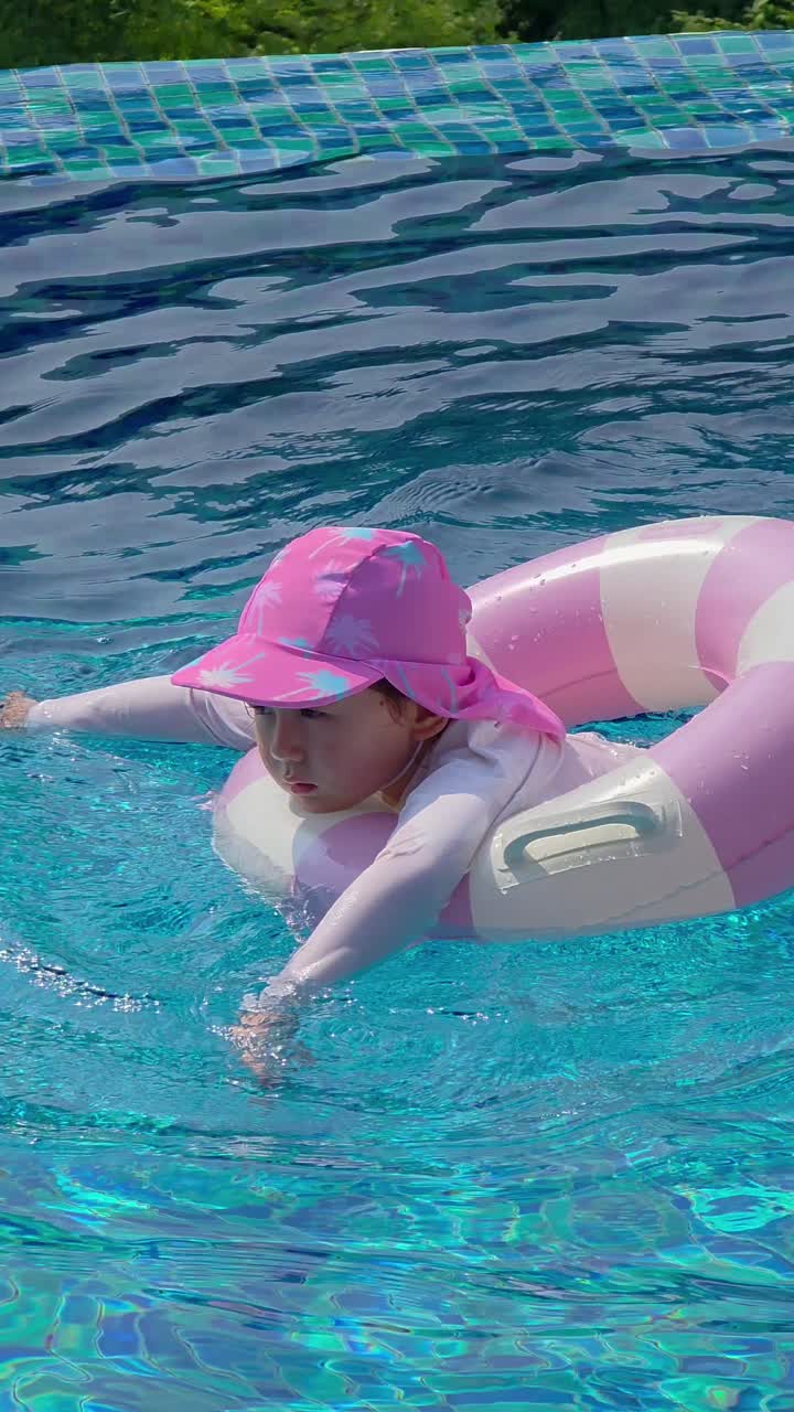 A young child swims in a clear blue pool using a pink and white float ring, wearing a pink sun hat and a long-sleeve rash guard for sun protection