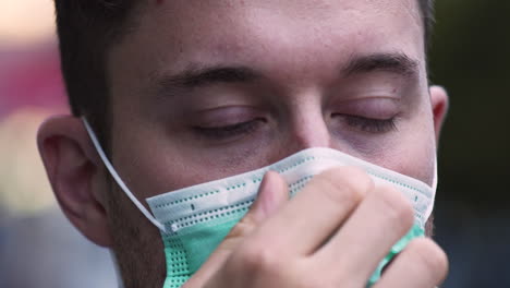 um jovem com olhos azuis brilhantes e barba por fazer coloca uma máscara protetora verde covid-19 em público, ajustando-a no nariz e no rosto, olhando diretamente para a câmera, close-up estático 4k shot