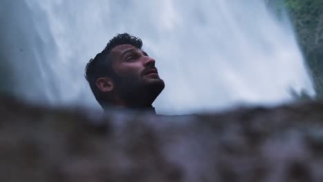 Young-male-slow-motion-gazing-up,-sitting-in-front-a-waterfall-in-peace,-closeup