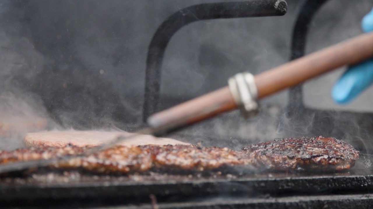 Cook flips sizzling meat patties on smoky grill using spatula, wearing blue gloves, outdoors