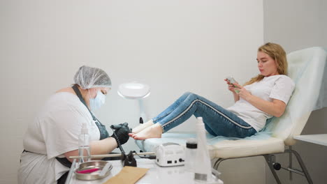 beautician wearing gloves and mask filing client toenails using electric filer under bright lamp while client relaxes on treatment chair using smartphone, with pedicure tools neatly arranged on table