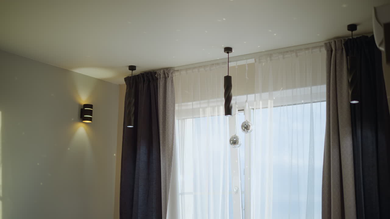 Modern living room interior with ceiling pendant lamps, glowing wall light, sheer curtains, dark drapes, daylight window view and hanging decorative disco balls creating cozy warm home atmosphere