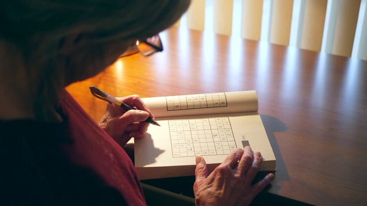 Donna caucasica di mezza età con mani segnate dal tempo che risolve un libro di giochi numerici per la lucidità mentale, per mantenersi mentalmente agile e in salute.
