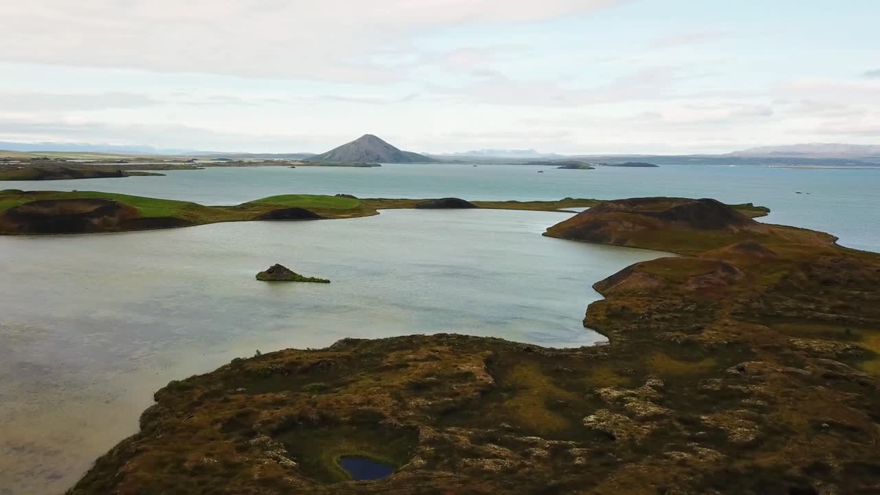 Panoramic view of volcanic craters surrounded by a serene lake at Skútustaðagígar, Iceland, with distant mountains under a soft, cloudy sky, capturing the unique beauty of the Icelandic landscape