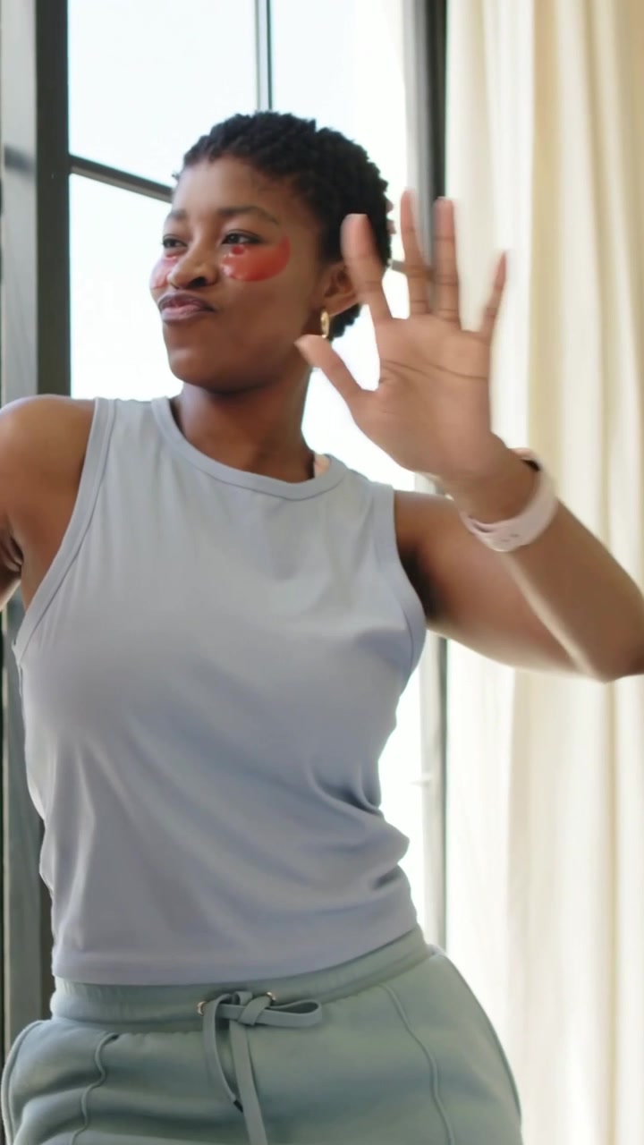 Vertical video: Extending right arm, woman turning toward cam near window, posing with eye patches