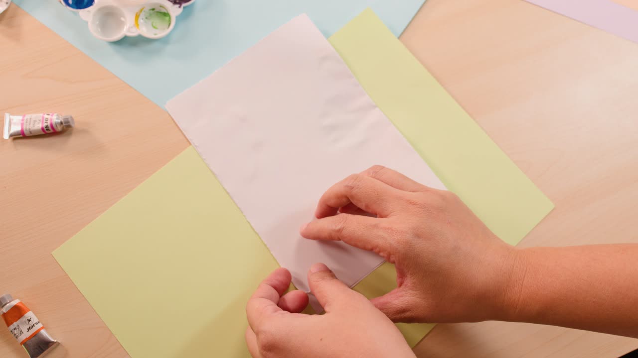 Two children fold paper to reveal colorful symmetrical paint blot artwork under soft indoor lighting