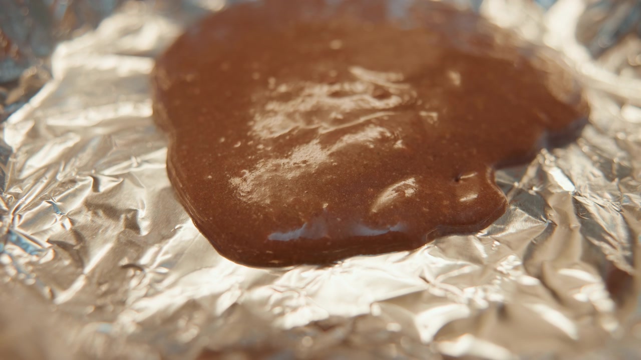 Primo piano di impasto al cioccolato su carta stagnola, miscela marrone lucida che riposa su alluminio increspato, consistenza densa che cattura la calda luce della cucina, evoca la preparazione di un dolce fatto in casa e la ricchezza tattile.