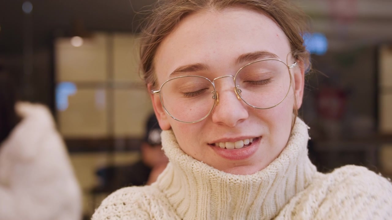Young woman wearing knit sweater, smiling with eyes closed in warm indoor cafe, adjusting turtleneck and playing with oversized knit, soft lighting, eyeglasses, gentle relaxed mood, candid lifestyle