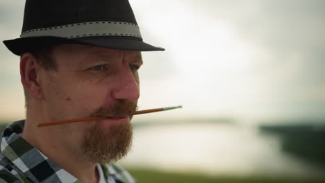 close-up of a man with a brush in his mouth, wearing a hat and checkered shirt, deeply focused on an unseen task. the cloudy sky in the background adds to the contemplative atmosphere