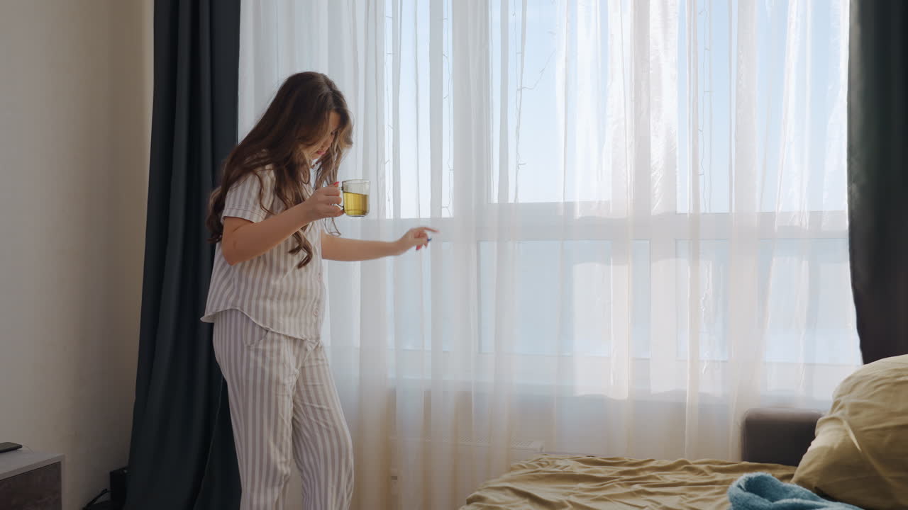 Woman Sipping Coffee By Window, Bright Bedroom Light And Cozy Bedspread, Soft Curtains, Relaxed Morning Routine, Steaming Mug Held Near Face, Barefoot On Floor, Casual Pajamas And Calm Atmosphere