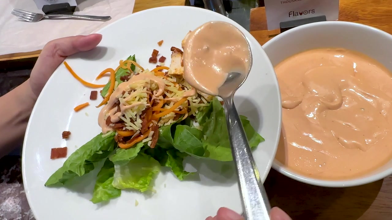 A person spoons creamy orange dressing over a salad with lettuce, bacon, and cheese