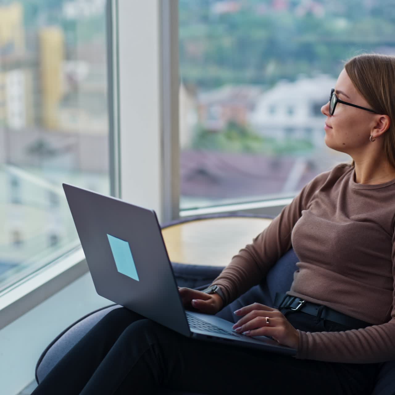 Good-looking blonde lady working on laptop sitting in bean bag chair. Lady distracts from work looking into big panoramic window she's sitting by