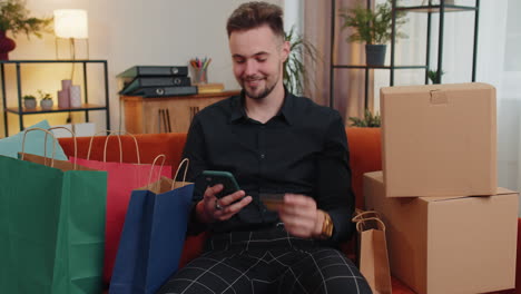 happy shopaholic young guy sitting with shopping bags making online payment with credit card