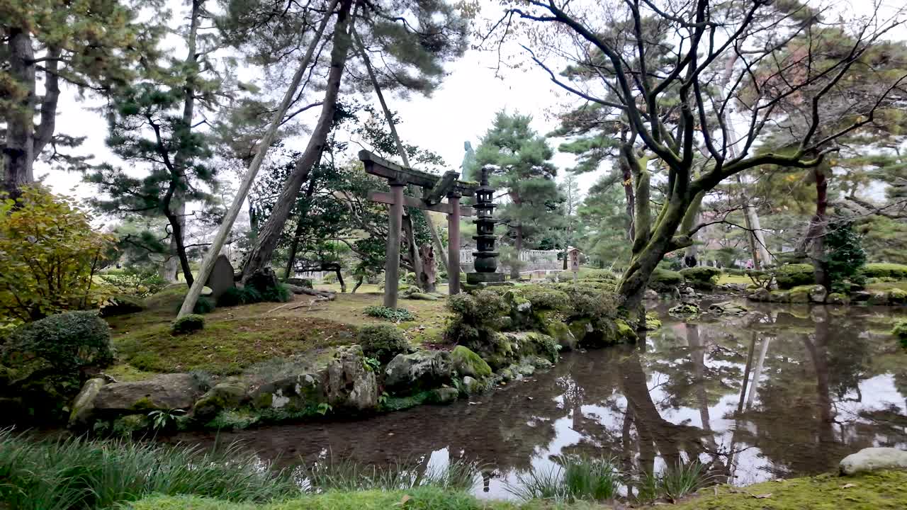 Traditional torii gate and stone lantern stand beside pond reflecting trees in Kenrokuen Garden, Kanazawa, Japan