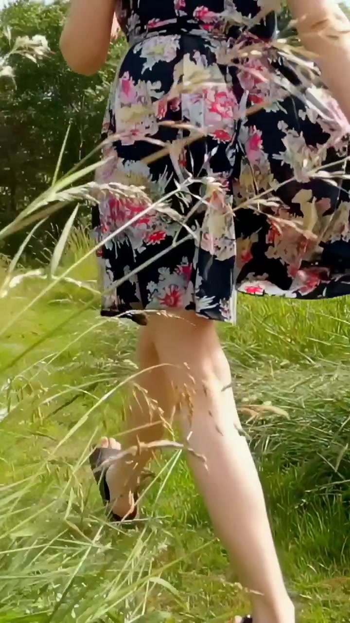 Vertikal video: Træder ind på sti, kvinde går mod træer i sort blomstret kjole og brune sandaler.