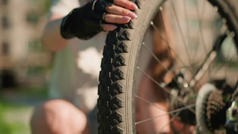 close-up of rear bicycle tire with knobby tread pattern, inspected for air pressure by person wearing black gloves in sunny park setting, background softly blurred