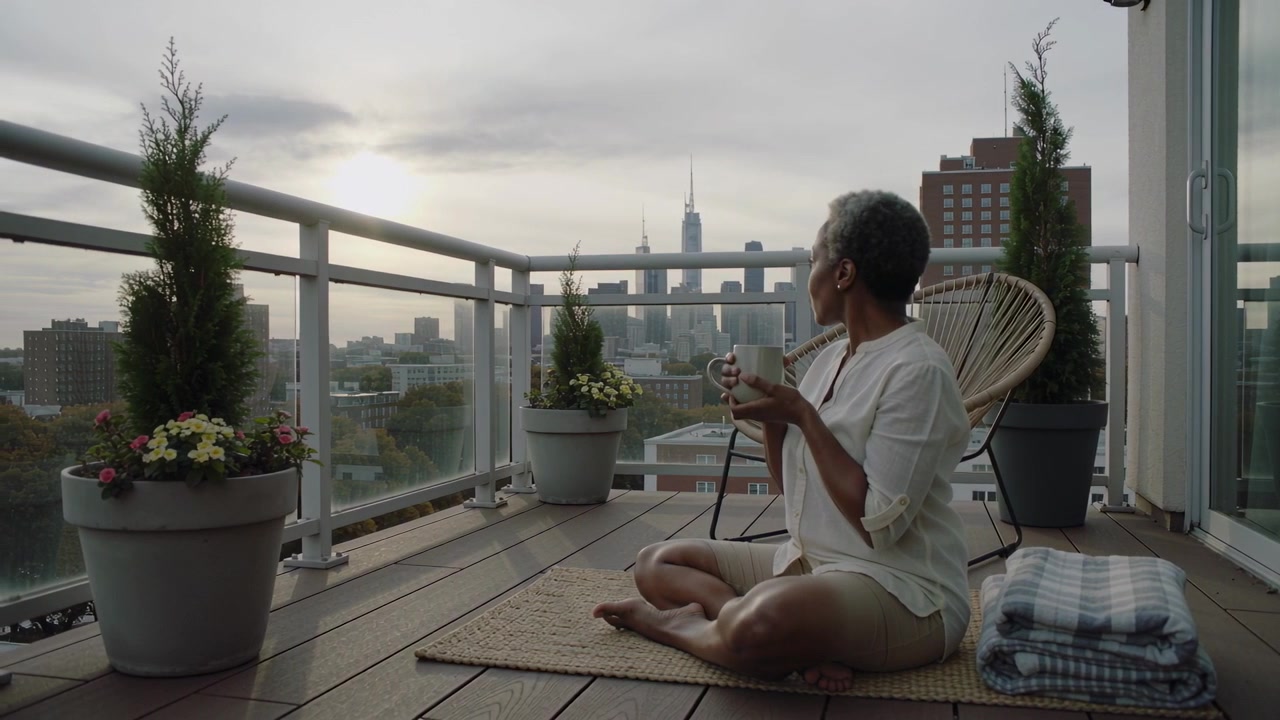 Mulher de cabelos grisalhos vestindo camisa, segurando uma caneca e observando o horizonte na varanda enquanto o sol ilumina, com espaço para texto