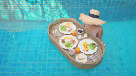 high angle view of a woman with a served breakfast on a floating tray or basket on the surface of a swimming pool