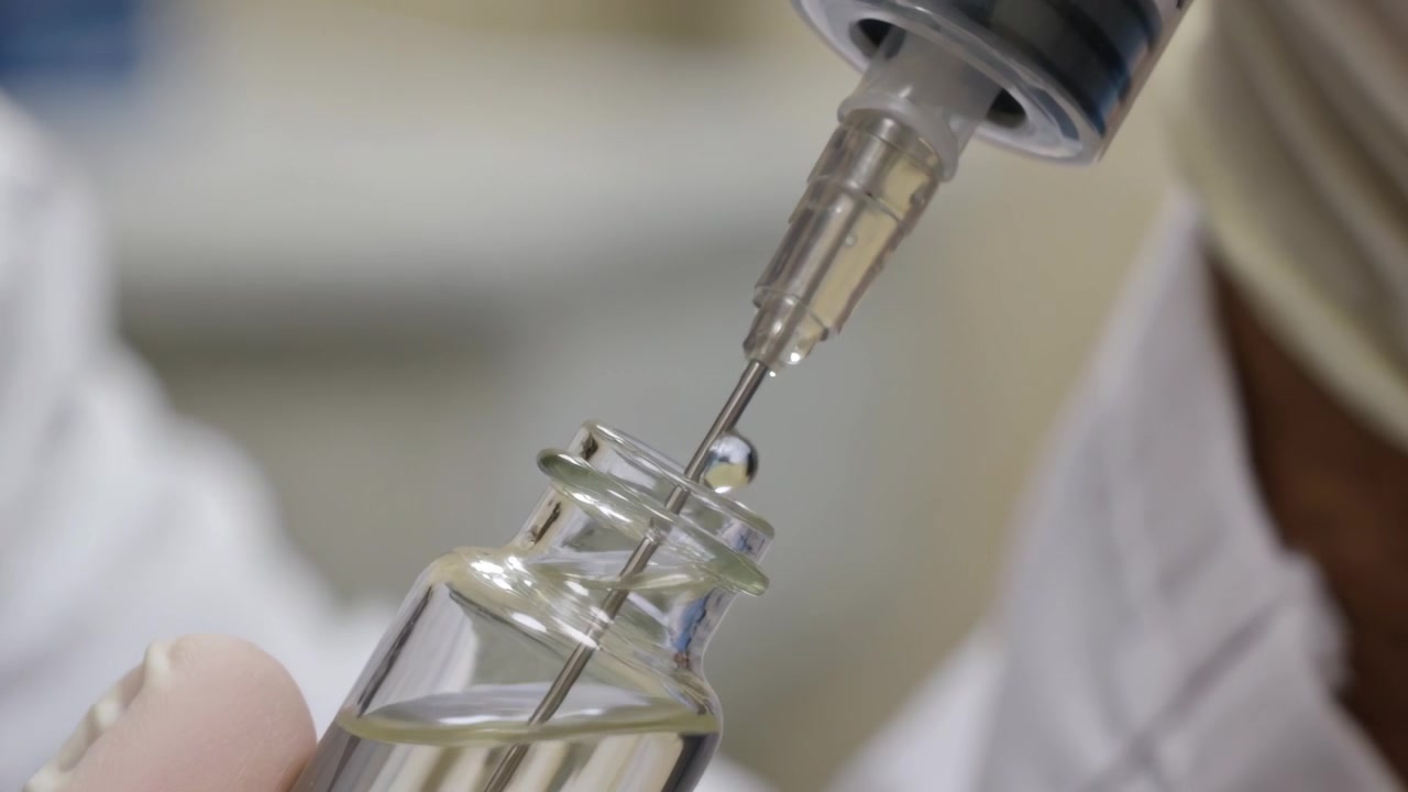 Tecnico di laboratorio in camice e guanti che fora il collo di una fiala, riempiendo una siringa al banco.