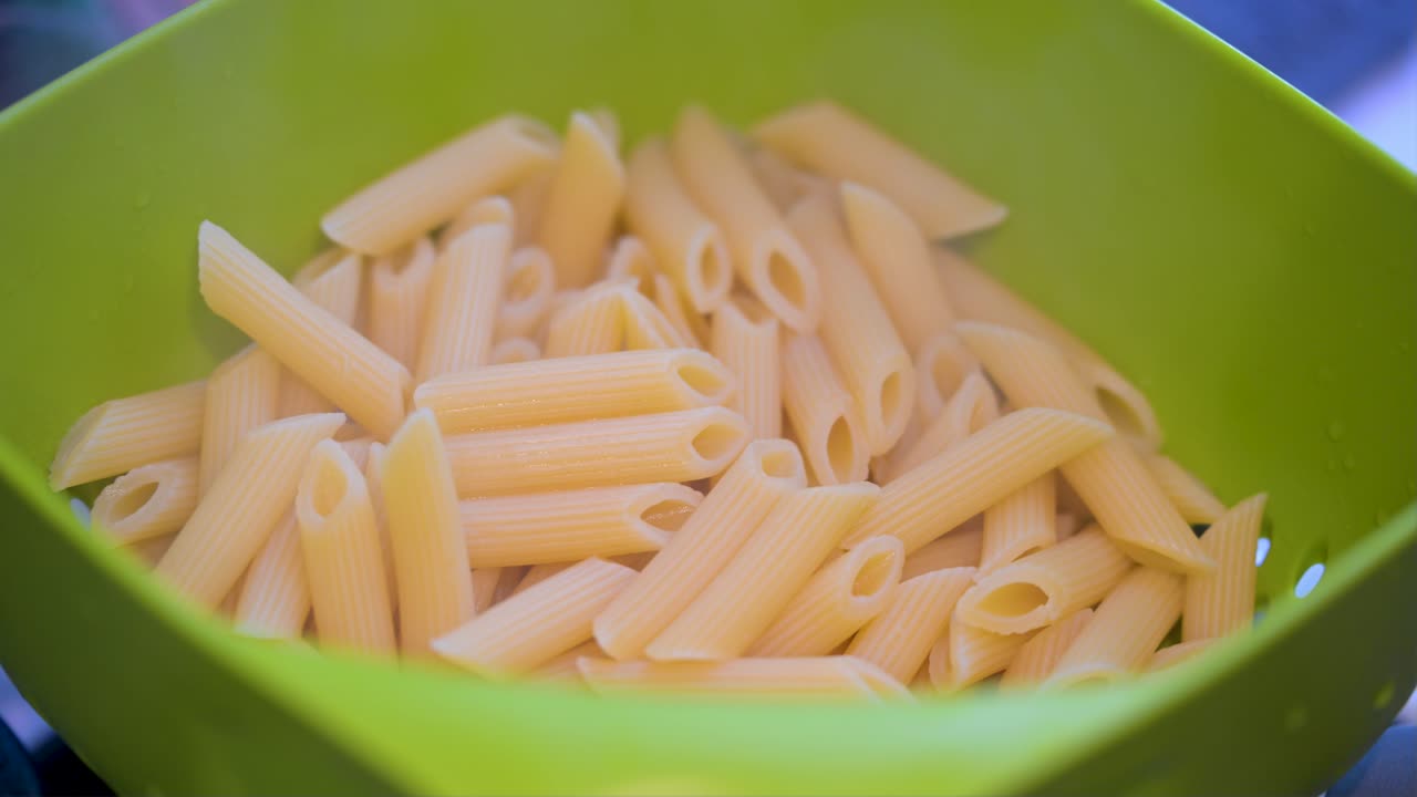 Freshly boiled penne pasta is drained in a colander in a kitchen. Steam gently rises from the hot pasta, capturing a key final step in the cooking process