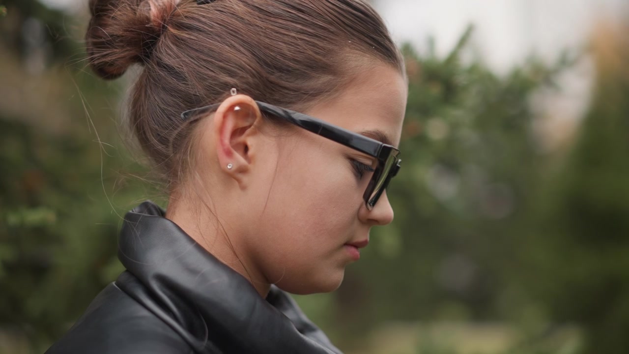Close up of young woman with brown hair in bun wearing black sunglasses and earrings dressed in leather jacket. scene is outdoors during daylight hours with greenery visible out of focus