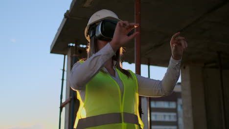 Eine-Bauingenieurin-Steht-Bei-Sonnenuntergang-Mit-VR-Brille-Auf-Dem-Dach-Eines-Gebäudes-Und-Bewegt-Ihre-Hände-Mithilfe-Der-Schnittstelle-Der-Zukunft.-Futuristischer-Ingenieur-Der-Zukunft.-Die-Ansicht-Von-Hinten.