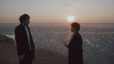 couple smoking and speaking on shore of frozen lake at sunset