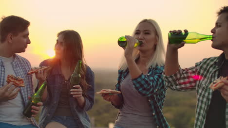 two loving couples drinks beer and eats hot pizza on the roof. they are smile and enjoy the time at the sunset. girl modestly looks at her boyfriend