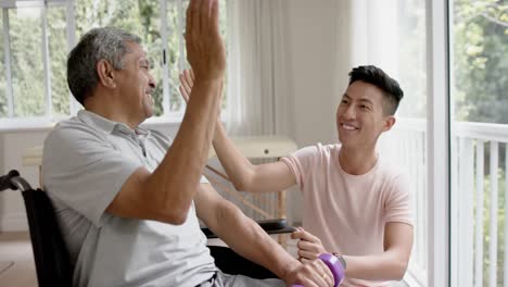 diverse male physiotherapist and senior man in wheelchair with dumbbells high fiving, in slow motion