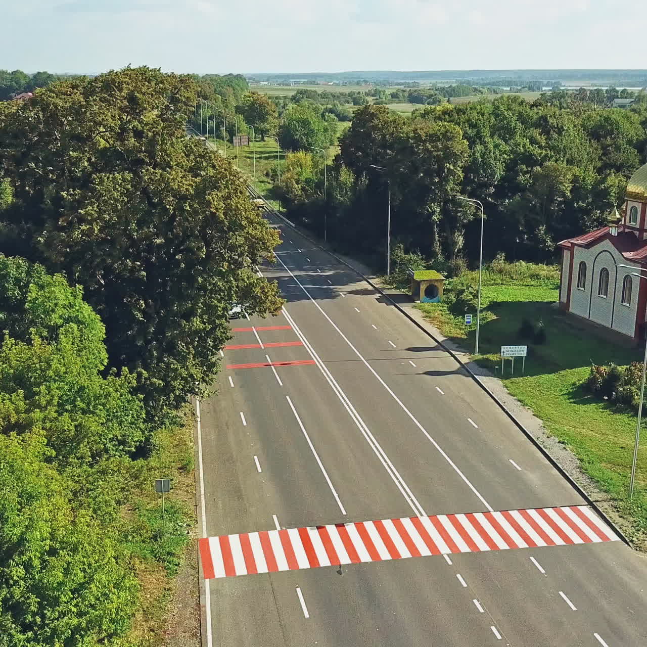 Aerial view of a new asphalt road. Flight over the highway surrounded by green trees in summer. Beautiful church near the road in the countryside.
