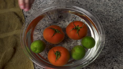 hands remove green bell peppers, red tomatoes, and green lime from glass bowl with water