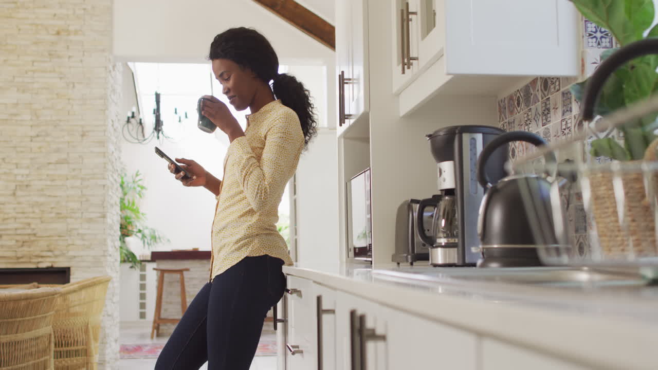Video of african american woman drinking coffee and using smartphone in kitchen