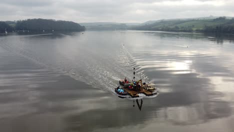 a small workboat tug pushing a barge with a crane on through the frame with a still river and countryside in the background