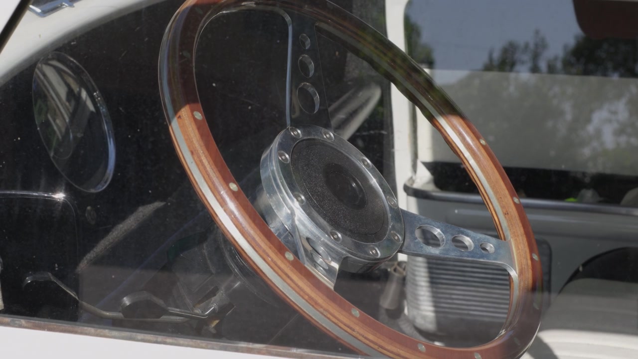 Detaillierte Nahaufnahme durch ein Fenster, die ein Retro-Holzlenkrad und Armaturenbrett in einem sonnendurchfluteten Oldtimer-Innenraum zeigt, keine Personen sichtbar