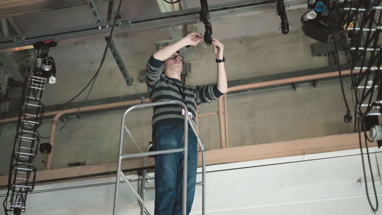 Person arbejder på hævet platform, justerer lys- eller kameraudstyr monteret på loftsgitter, med synlige kabler, iført sweater og briller, i et industrielt miljø, udfører vedligeholdelses- eller installationsopgave