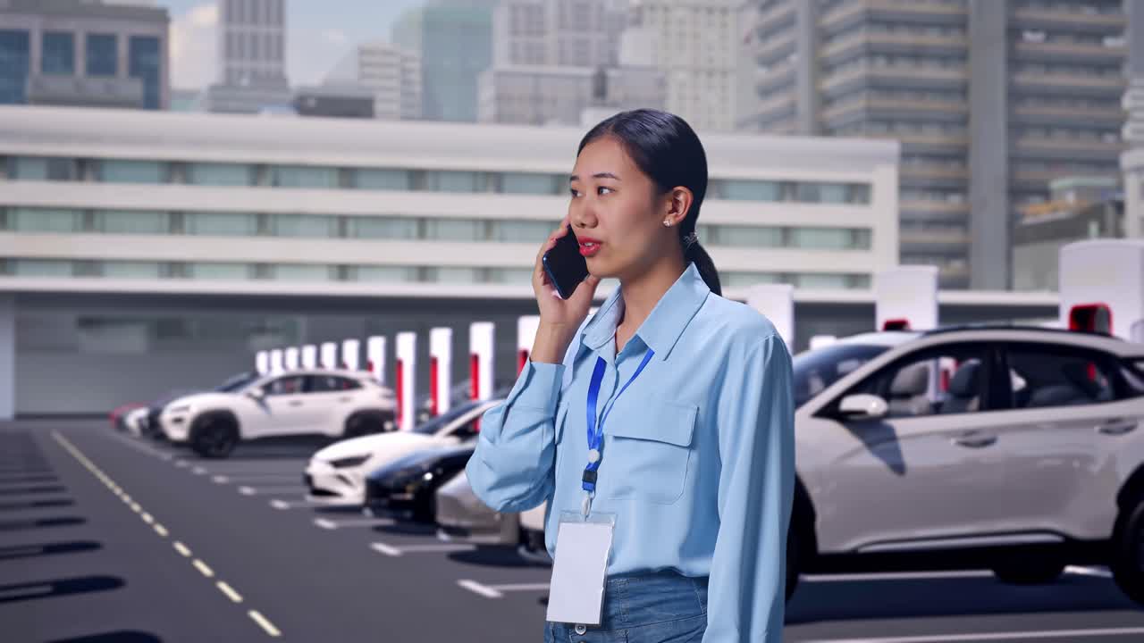 現代的な都市の充電ステーションで電気自動車が充電されている中、横顔のアジア人女性が電話を取り上げ、わずかな笑顔で話し始めた。