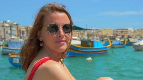 Close-Up-Of-A-Red-haired-Woman-In-Sunglasses-By-The-Sea-Port-Looking-Around-And-Into-The-Camera-In-Summer