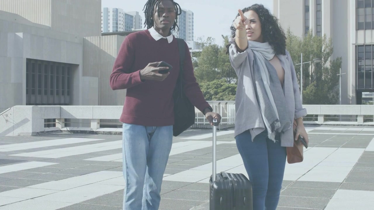 Traveling duo walking plaza woman pointing and pulling suitcase man checking phone confirming route